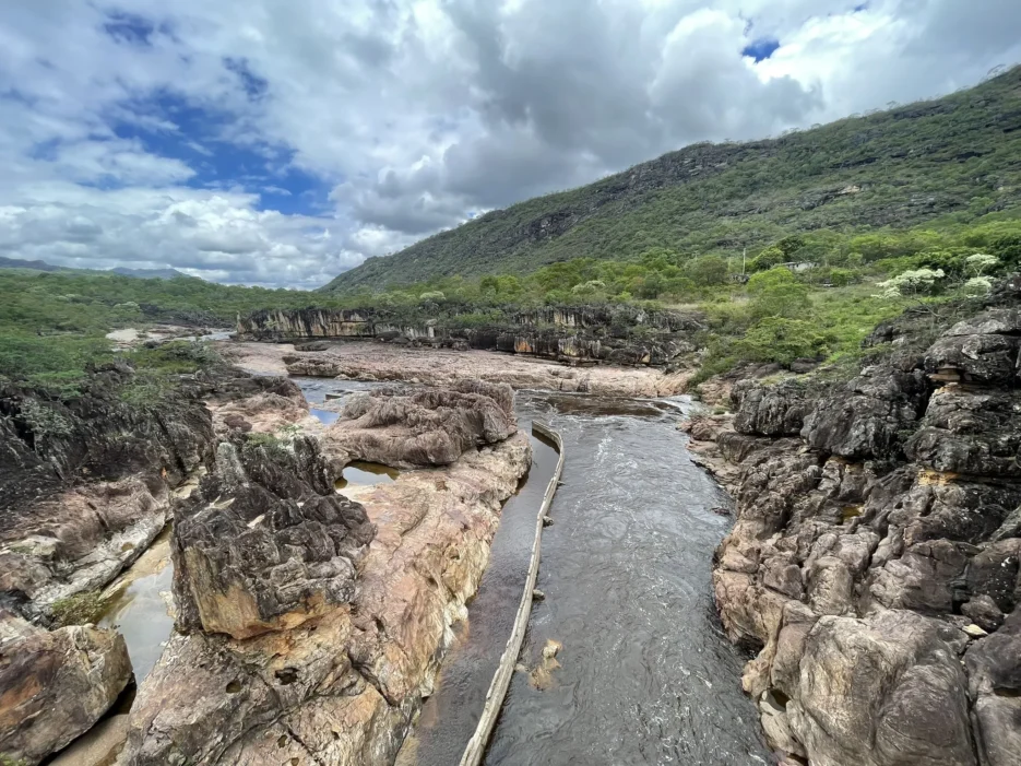 Rio atravessa formações rochosas cercadas por vegetação nativa e serras sob um céu nublado.