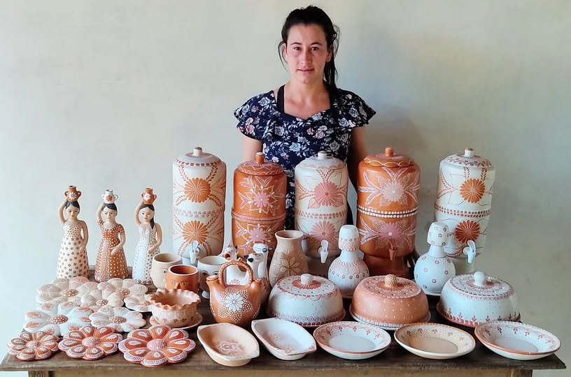 Artesã atrás de uma mesa com bonecas de barro, vasos, moringas e peças de cerâmica decoradas com motivos florais.