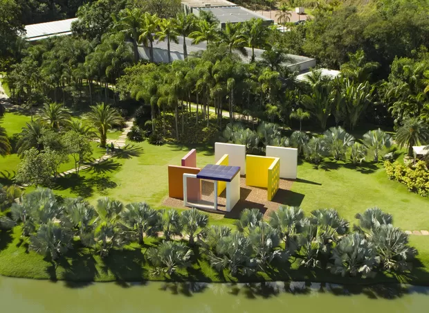 Vista aérea de uma instalação artística com estruturas coloridas, cercada por palmeiras e jardins do Instituto Inhotim, em Brumadinho.