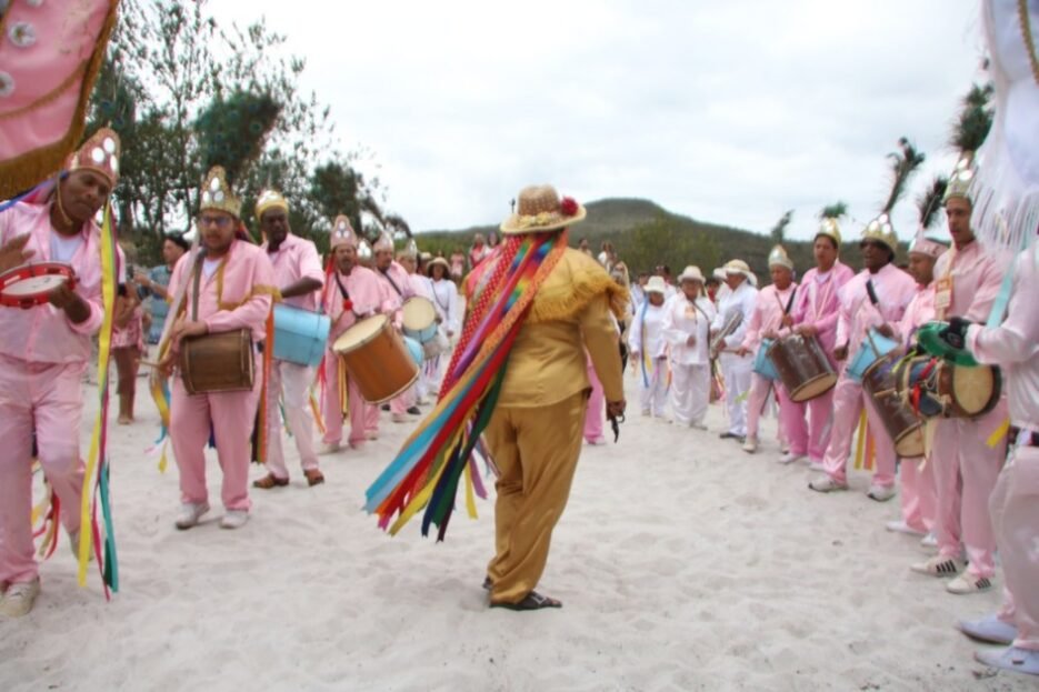 Grupo de participantes com roupas rosas e brancas toca tambores durante uma celebração popular, enquanto uma pessoa com fitas coloridas dança ao centro.